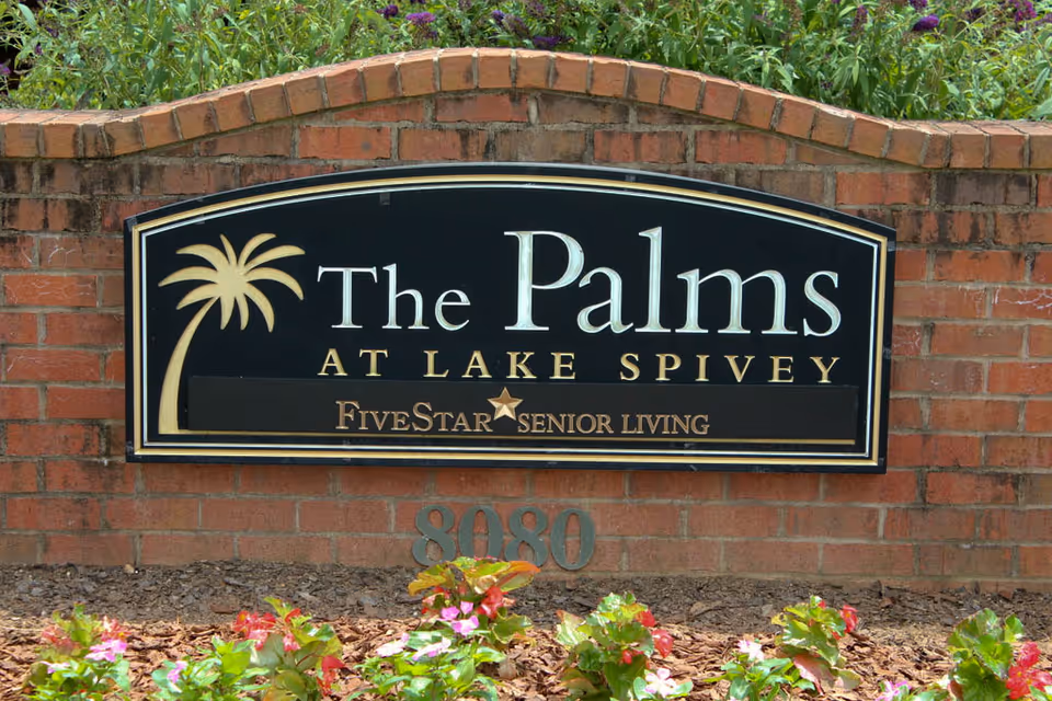 A black and gold sign mounted on a brick wall with flowers and greenery in front. The sign reads 'The Palms at Lake Spivey Five Star Senior Living' with a palm tree graphic on the left side. The number 8080 is displayed below the sign.