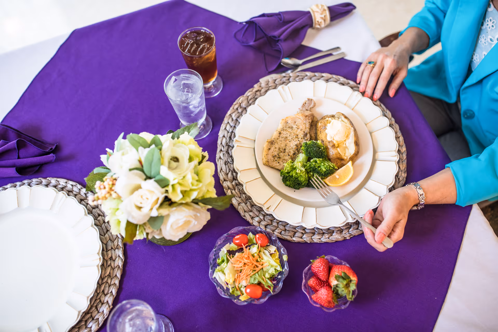 A person in a turquoise jacket is seated at a dining table with a purple tablecloth, eating a meal consisting of chicken, broccoli, a baked potato with sour cream, and a lemon wedge. The table is set with a floral centerpiece, a small bowl of salad, a bowl of strawberries, a glass of iced tea, and a glass of water.