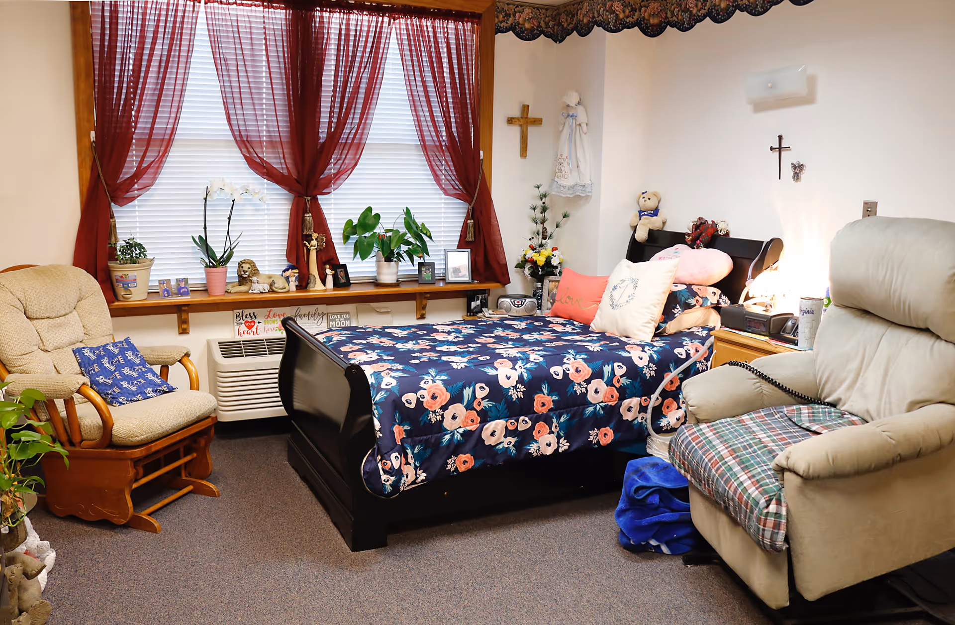 A cozy bedroom in a nursing and rehabilitation center featuring a bed with a floral comforter and multiple pillows, a beige recliner chair with a plaid cushion, a wooden rocking chair with a blue pillow, and a window with red curtains and white blinds. The room is decorated with plants, framed photos, a teddy bear, and religious crosses on the walls.