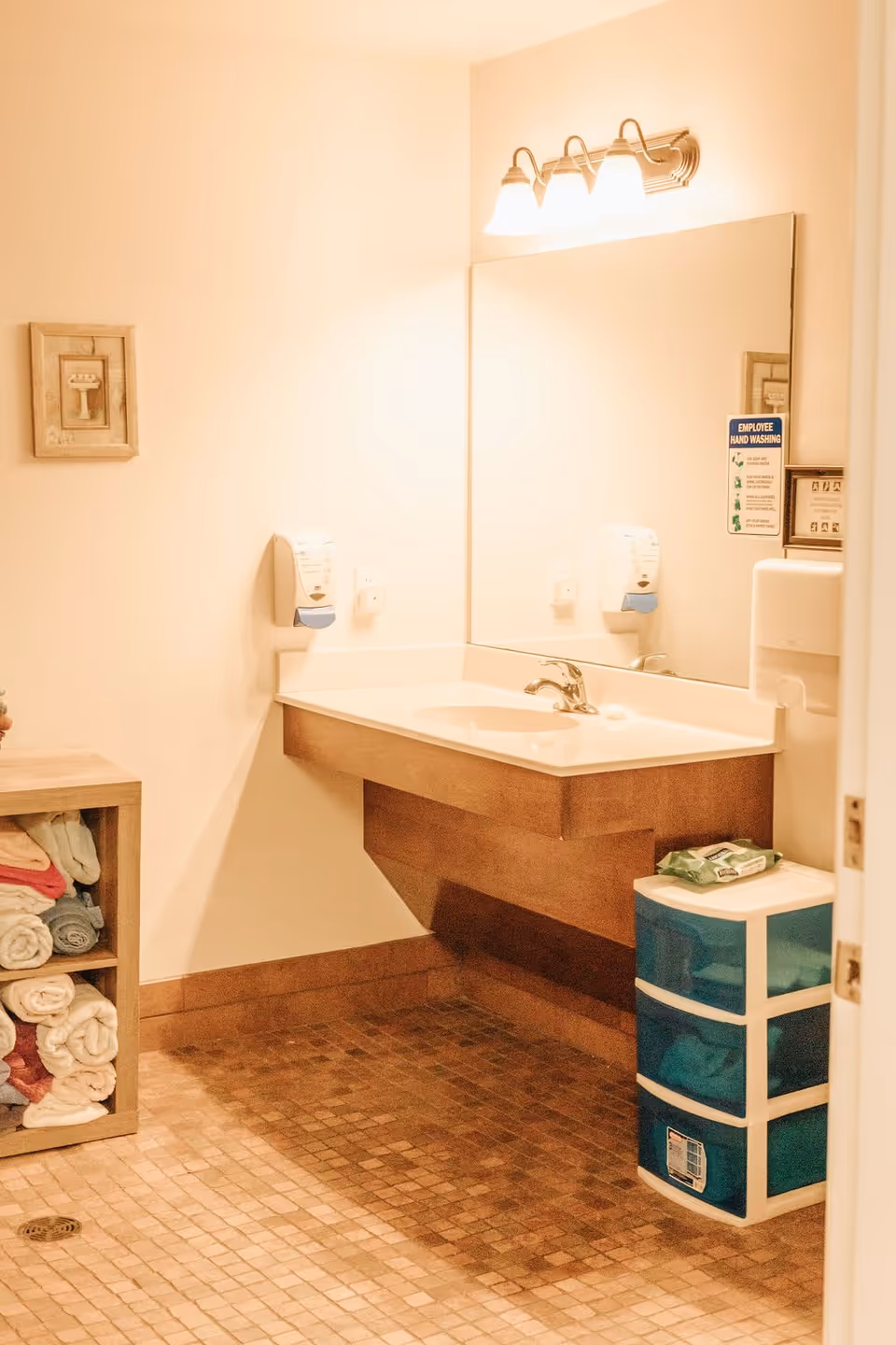 A clean bathroom area with a wall-mounted sink and faucet, a large mirror above the sink, two soap dispensers on the wall, a small shelving unit with neatly folded towels, and a plastic drawer unit with wipes on top. The floor is tiled with small square tiles, and there is a framed picture on the wall.