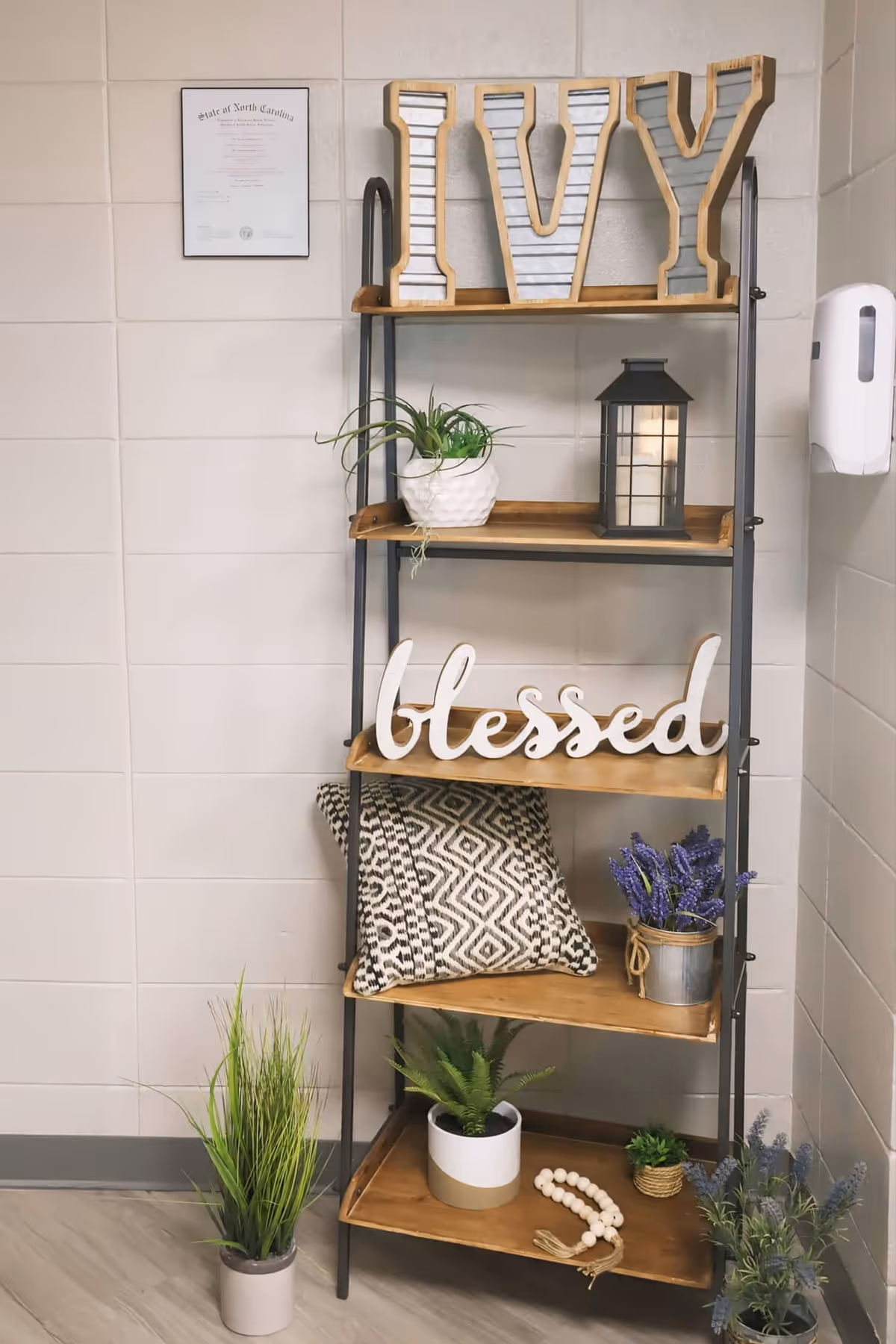 Decorative metal-and-wood shelving against a tiled interior wall holding large 'IVY' letters, a 'blessed' sign, potted plants, a lantern, patterned pillows and other decor.