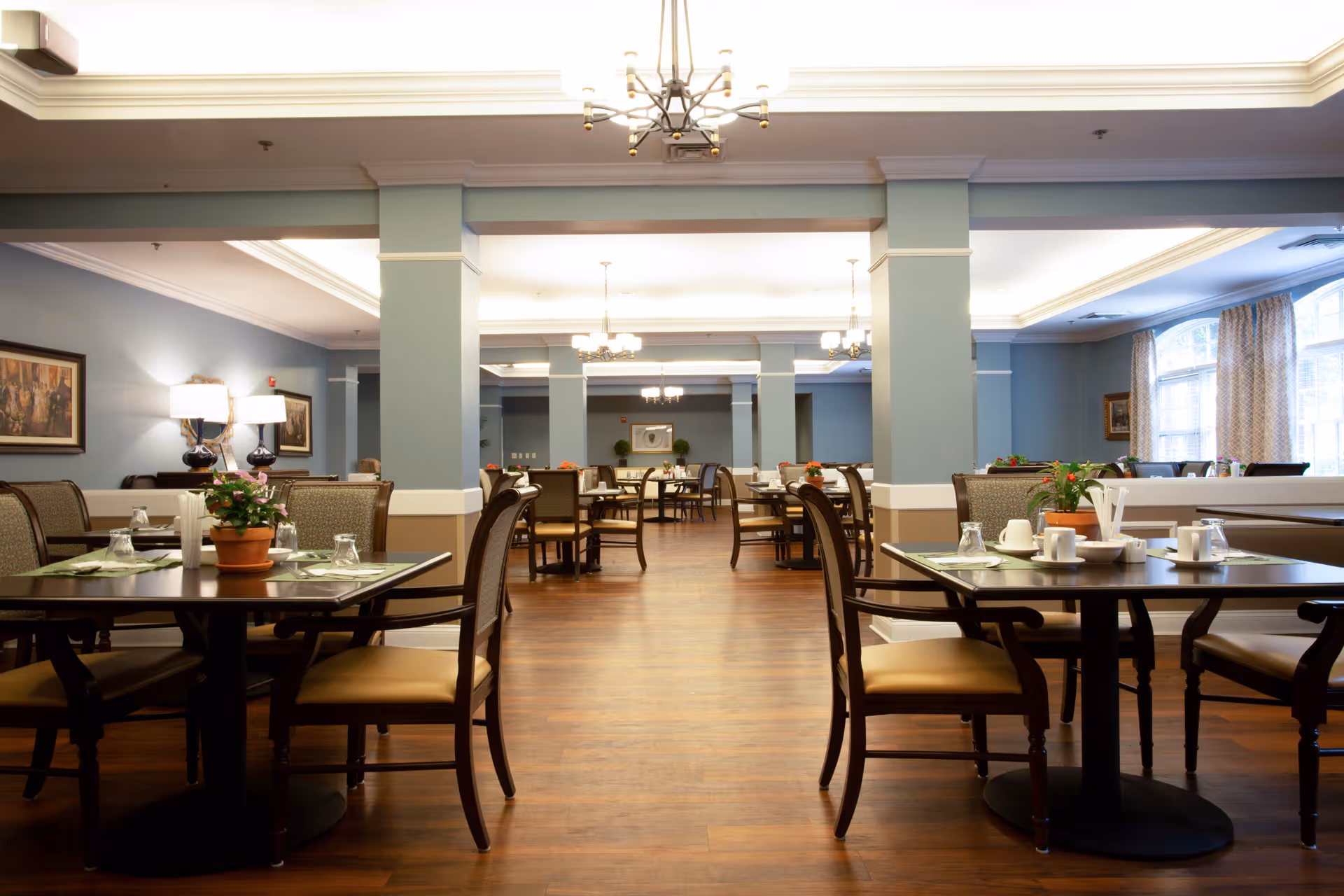A spacious dining room with multiple tables and chairs arranged neatly. Each table is set with placemats, cups, glasses, and small potted plants. The room features wooden flooring, soft blue walls, large windows with curtains, and elegant ceiling lights.