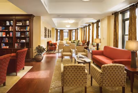 A spacious senior living lounge with red sofas, patterned chairs, tables, lamps, and a bookshelf.