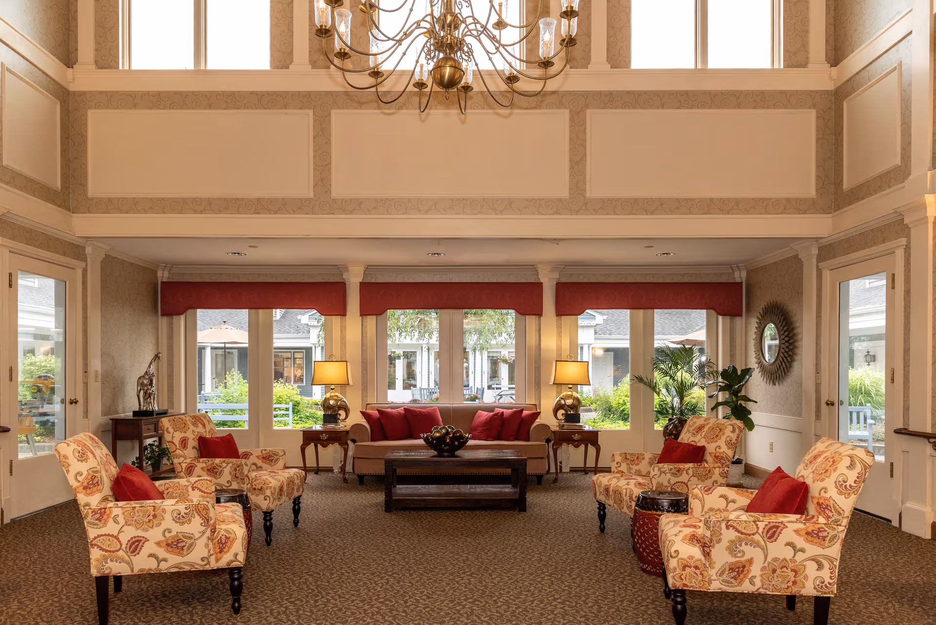 A spacious and elegant living room with high ceilings and large windows letting in natural light. The room features a beige sofa with red cushions, four patterned armchairs with red pillows, two side tables with lamps, a coffee table with a decorative bowl, and various plants and decorative items. The walls are light-colored with molding details, and a large chandelier hangs from the ceiling.