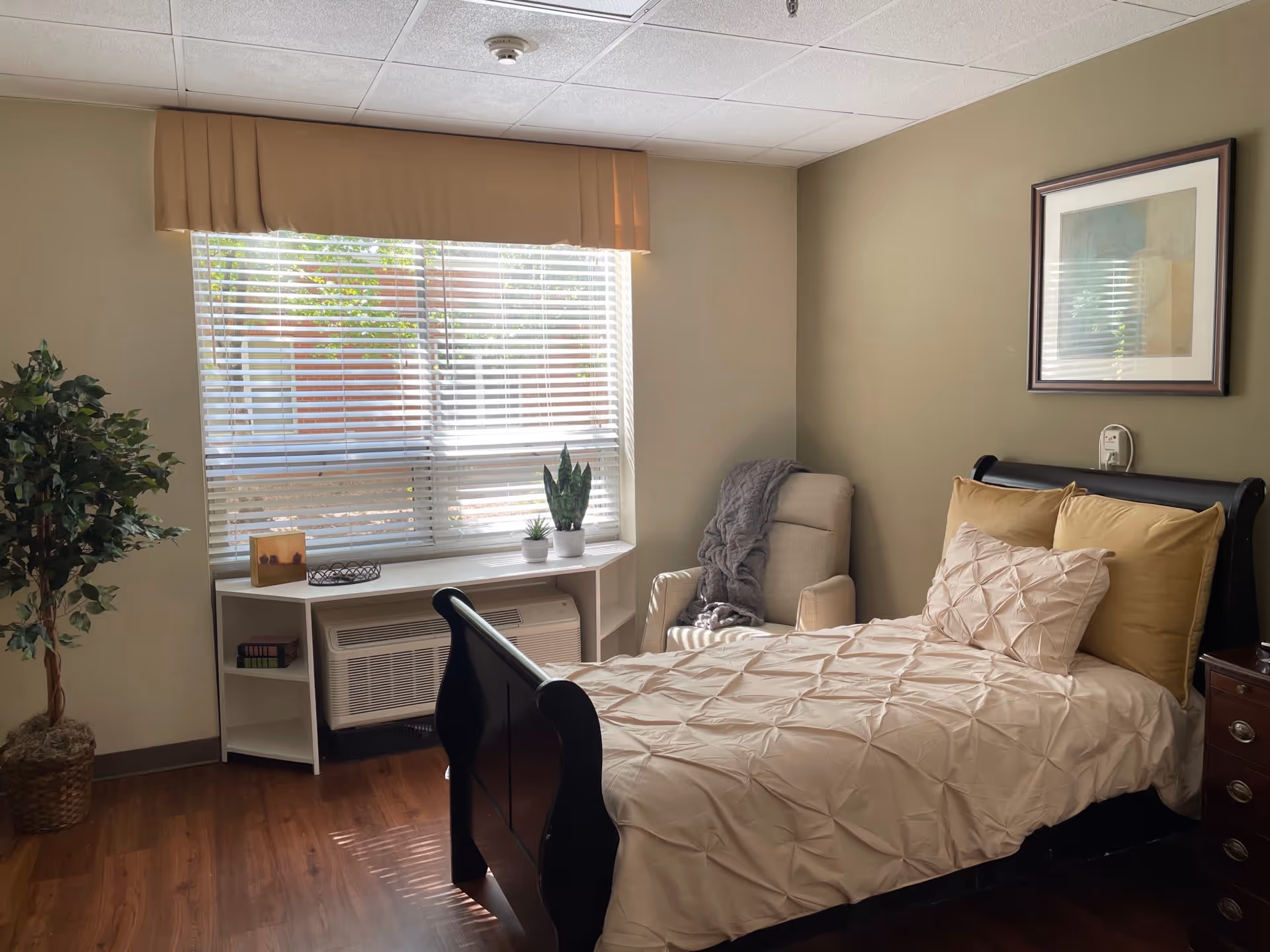 A cozy bedroom with a single bed featuring beige and cream bedding. Next to the bed is a comfortable armchair with a gray throw blanket. A large window with blinds and a beige valance lets in natural light. Below the window is a white shelf holding two small potted plants and a framed picture. A tall potted plant is in the corner, and a framed abstract artwork hangs on the wall above the bed.