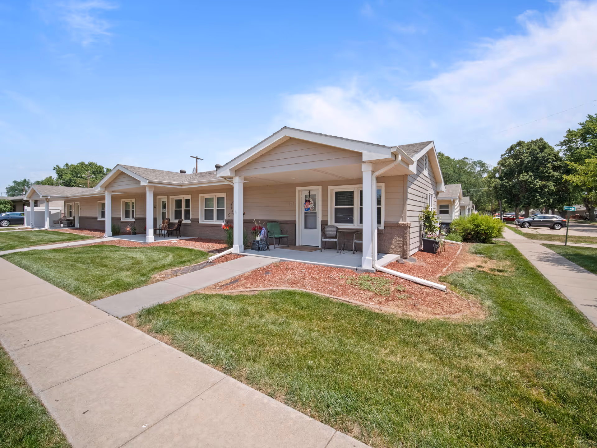 Exterior view of a single-story residential building with beige siding and white trim, featuring covered porches with chairs and small tables. The building is surrounded by well-maintained green lawns, sidewalks, and some landscaping with mulch and plants. The sky is clear and blue.