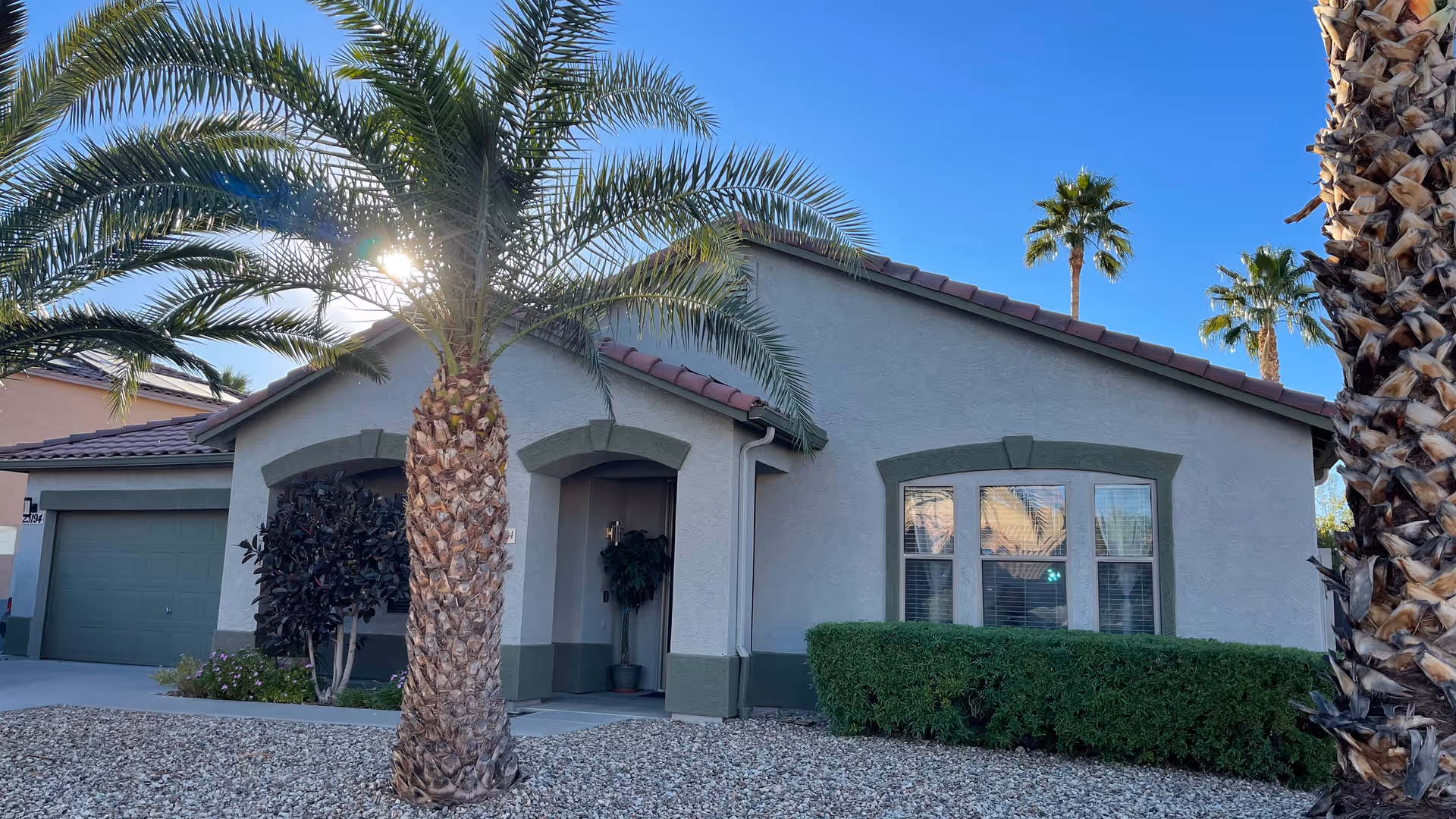 Exterior view of a single-story house with a tiled roof, a garage, and a front entrance partially shaded by palm trees and bushes under a clear blue sky.