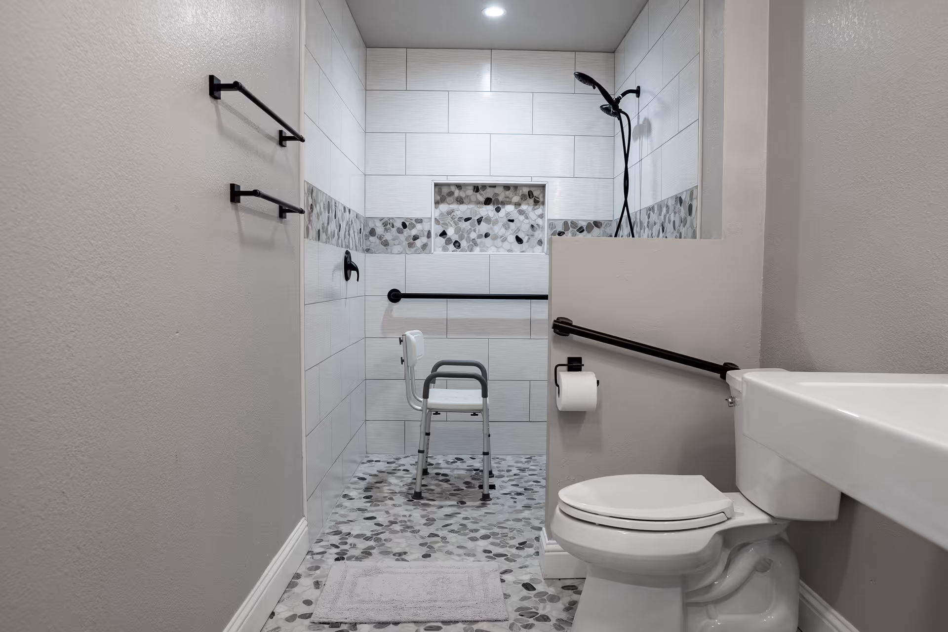 Accessible bathroom with a walk-in tiled shower featuring a shower chair and grab bars, plus a toilet and sink.