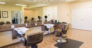 Interior view of a hair salon area in a retirement living facility featuring two salon chairs in front of sinks and a large mirror along the wall. Various hair care products are placed on the counter, and the floor has a tiled pattern.