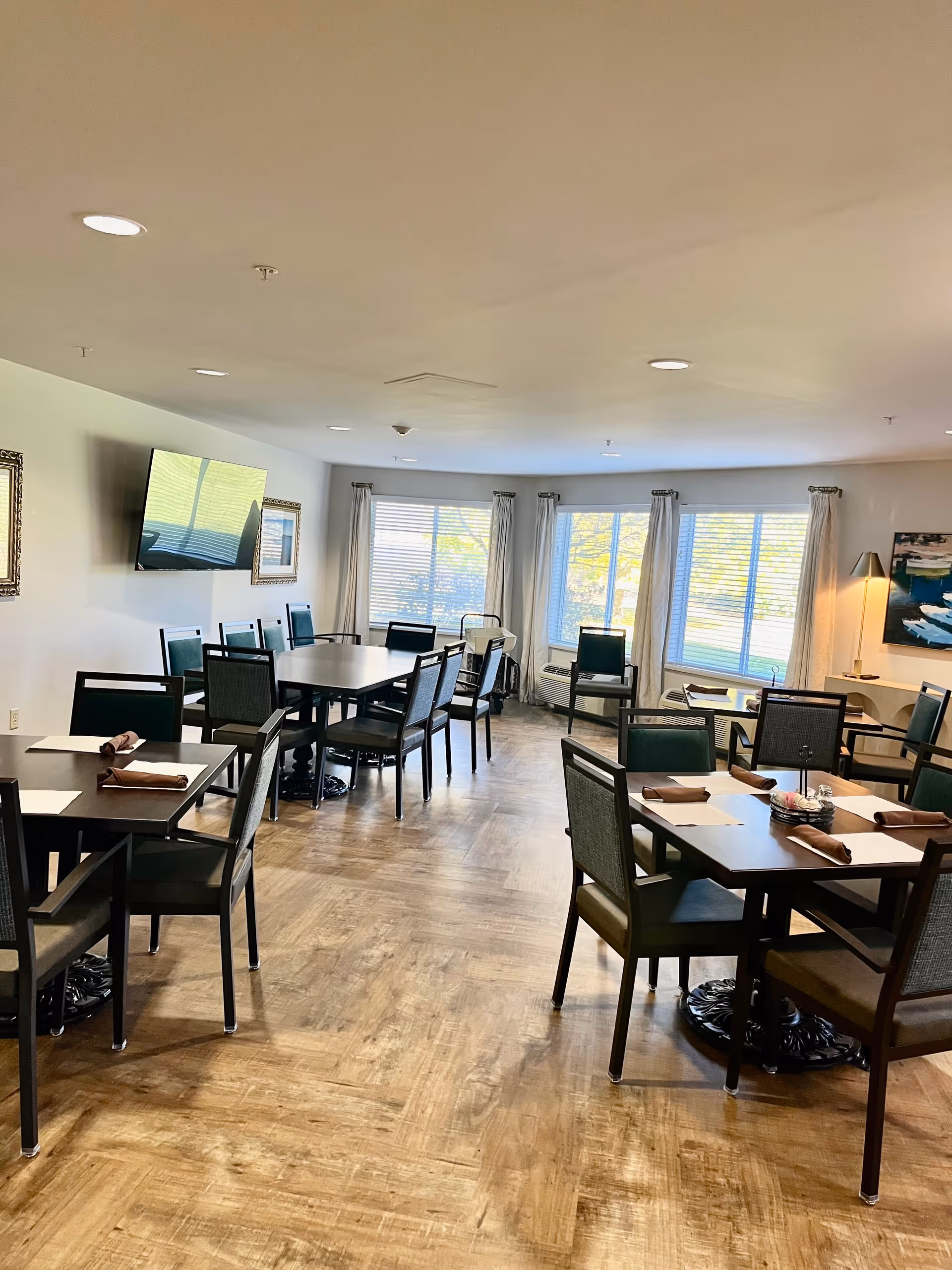 A bright dining room with several tables and chairs arranged neatly. Each table has place settings with napkins. Large windows with curtains allow natural light to fill the room. A flat-screen TV is mounted on the wall, and there is a floor lamp and framed artwork on the walls.