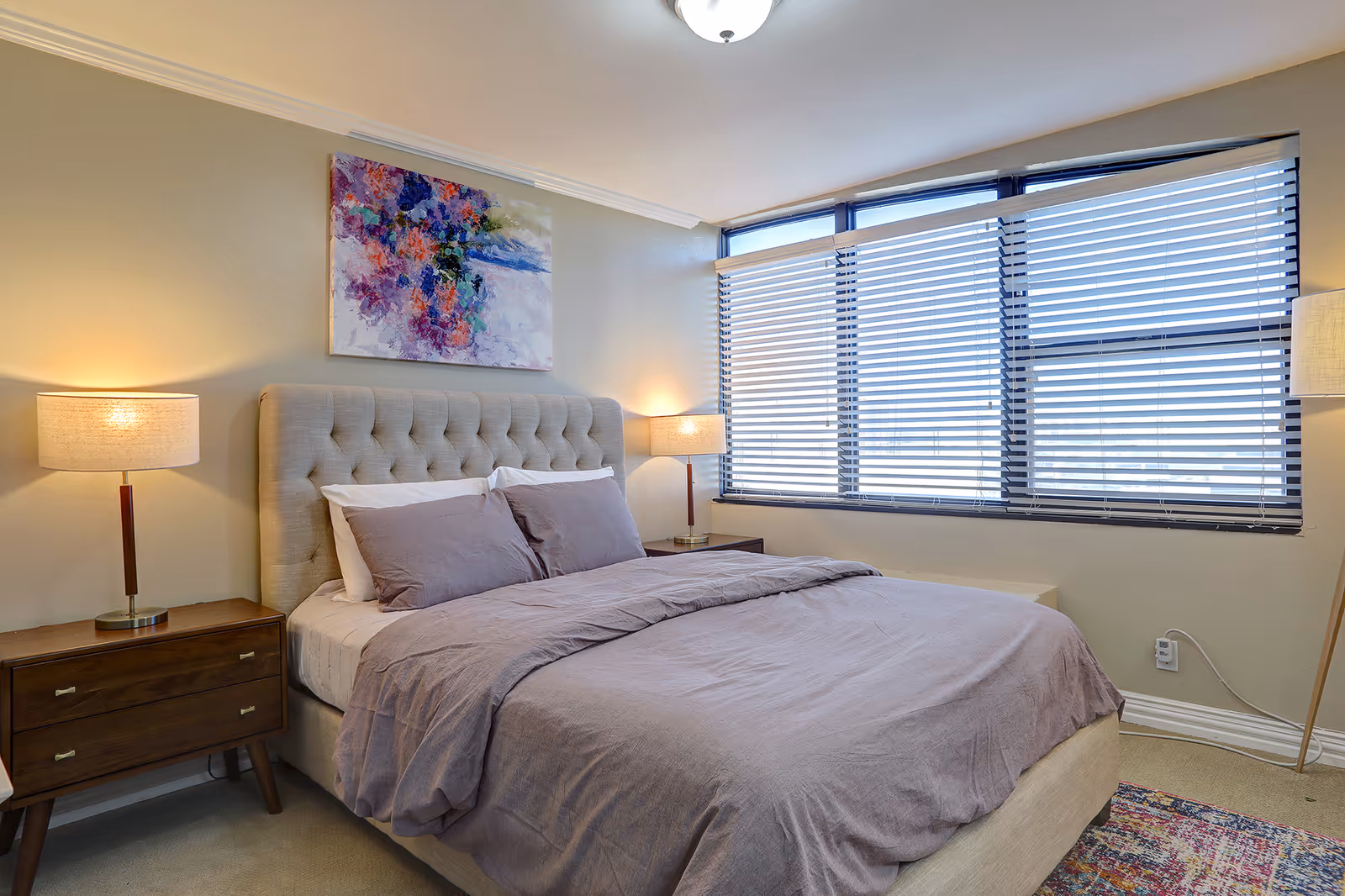 A cozy bedroom with a large bed featuring a tufted headboard, gray bedding, and two pillows. There are two wooden nightstands on either side of the bed, each with a lit table lamp. A colorful abstract painting hangs above the bed. Large windows with horizontal blinds allow natural light to fill the room. A patterned rug is partially visible on the floor.