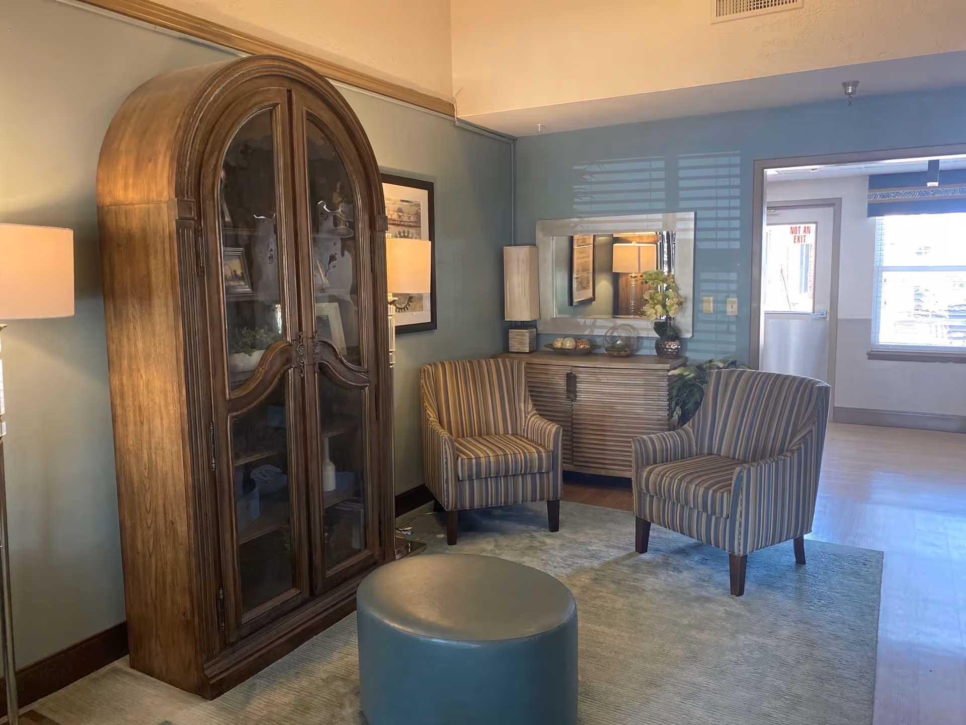 A cozy sitting area in a healthcare facility featuring two striped armchairs, a round blue ottoman, a wooden cabinet with glass doors displaying decorative items, a sideboard with a mirror above it, and two lamps providing warm lighting. The walls are painted light blue, and there is a window and a door in the background.
