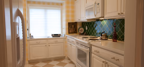 A bright kitchen with white cabinetry and appliances, including a refrigerator, stove, oven, and microwave. The backsplash features green tiles, and the walls have a yellow and white checkered pattern. There is a window with a white blind letting in natural light, and various kitchen items such as a kettle and decorative bottles are on the countertops.