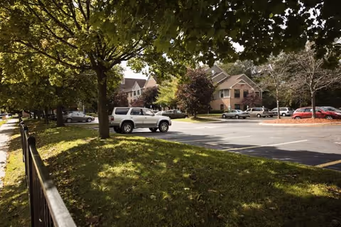 Parking lot with several parked cars next to a residential-style building surrounded by trees and greenery under a sunny sky.