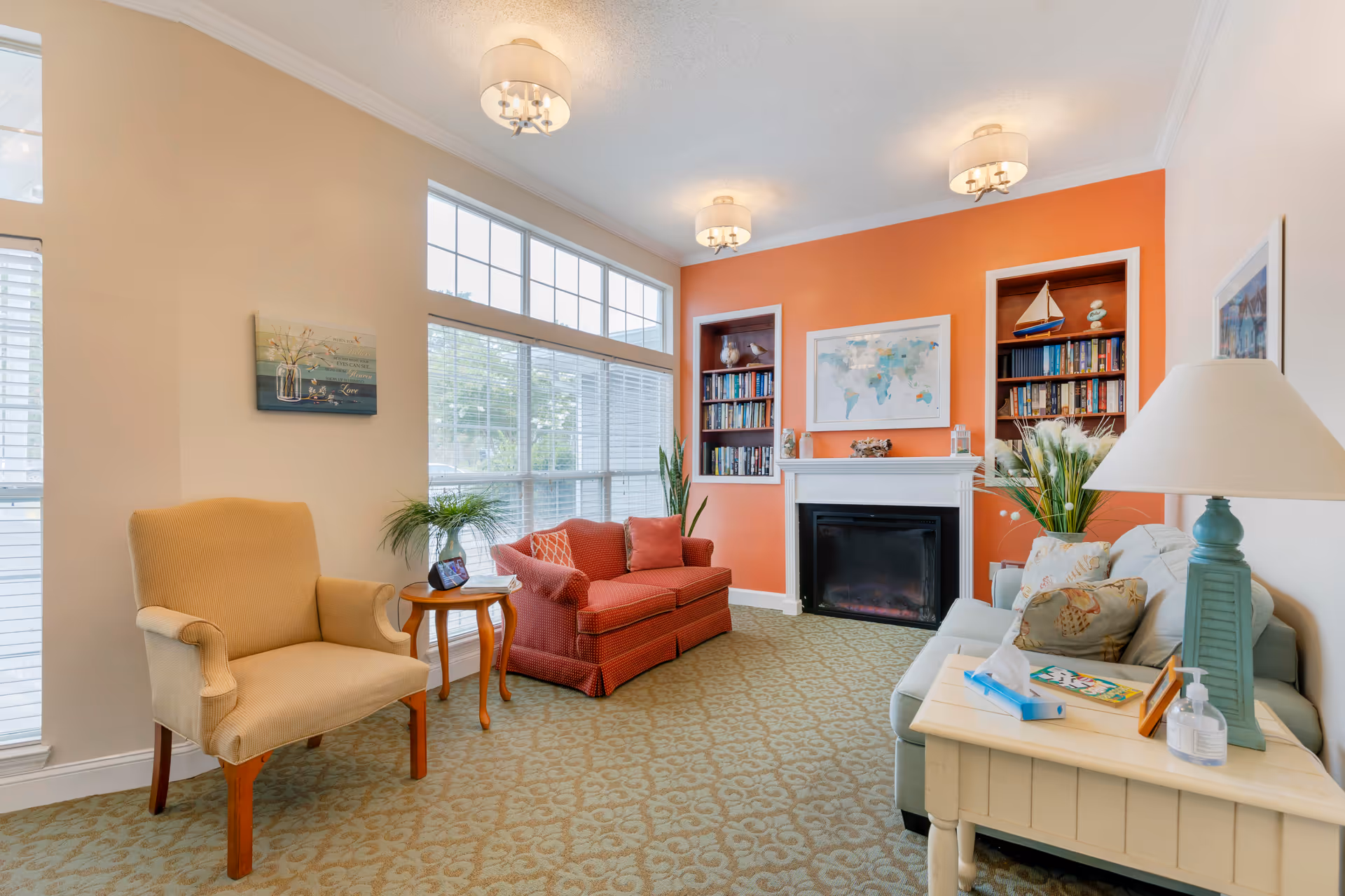 A cozy living room with a beige armchair, a red loveseat, and a light blue sofa. The room features a fireplace with a white mantel, flanked by built-in bookshelves filled with books and decorative items. Above the fireplace is a world map. Large windows with white blinds allow natural light to fill the space. The walls are painted in a combination of beige and orange, and the floor is covered with a patterned carpet. A side table with a lamp, tissues, and hand sanitizer is next to the sofa.