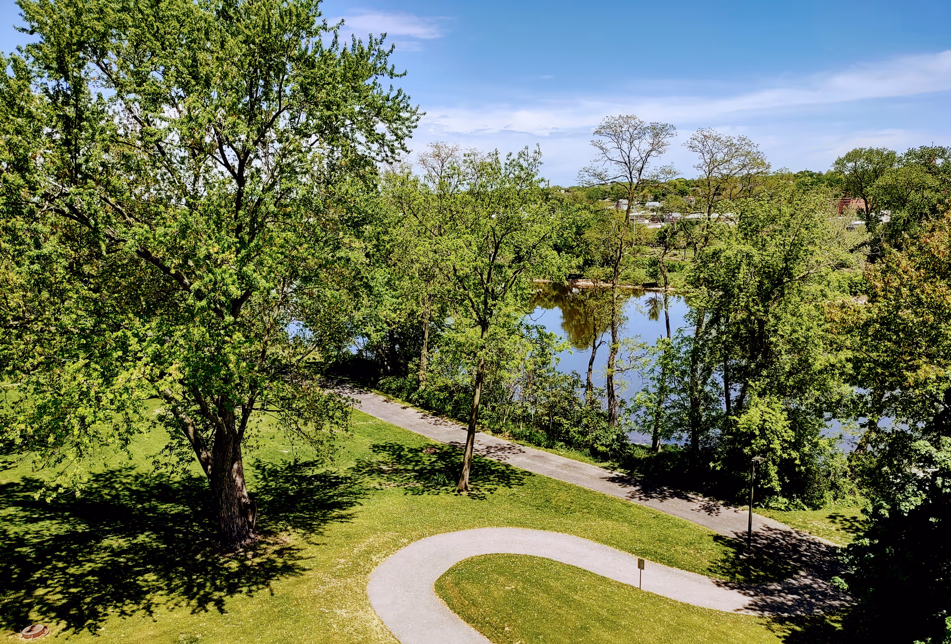 A scenic outdoor view at Plum Landing Retirement Community featuring a winding paved pathway surrounded by green grass and large leafy trees. In the background, there is a calm body of water partially visible through the trees under a clear blue sky.