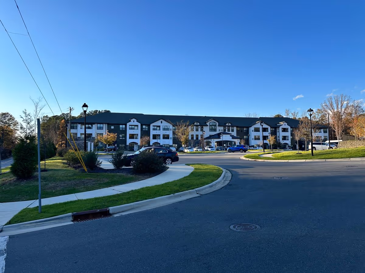 Front exterior of a three-story senior living building with a driveway, parked cars, and landscaped grounds under a clear blue sky.