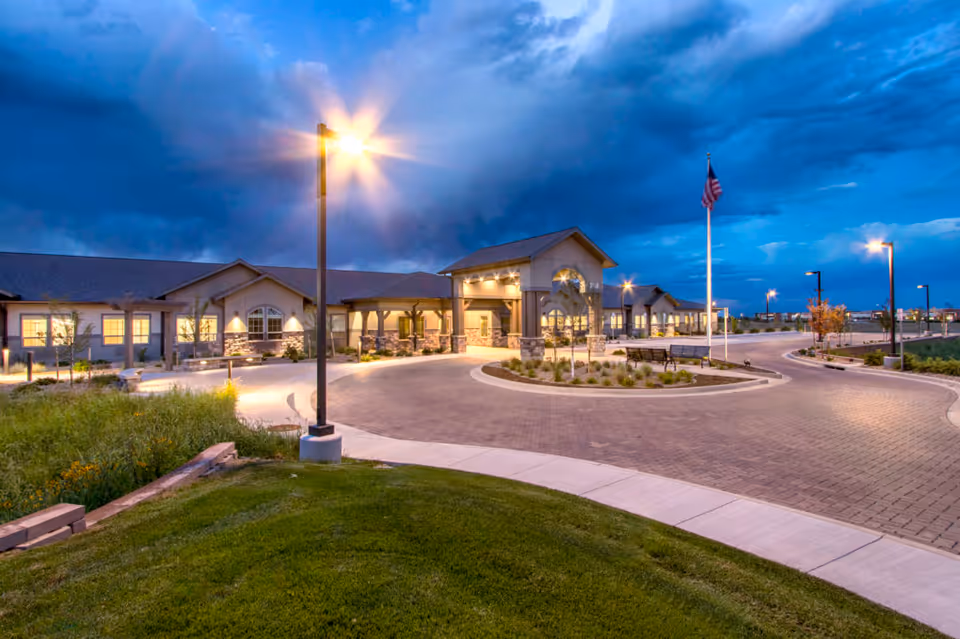 Front exterior of a memory care facility at dusk with a circular driveway, illuminated entrance, lampposts, and a flagpole.