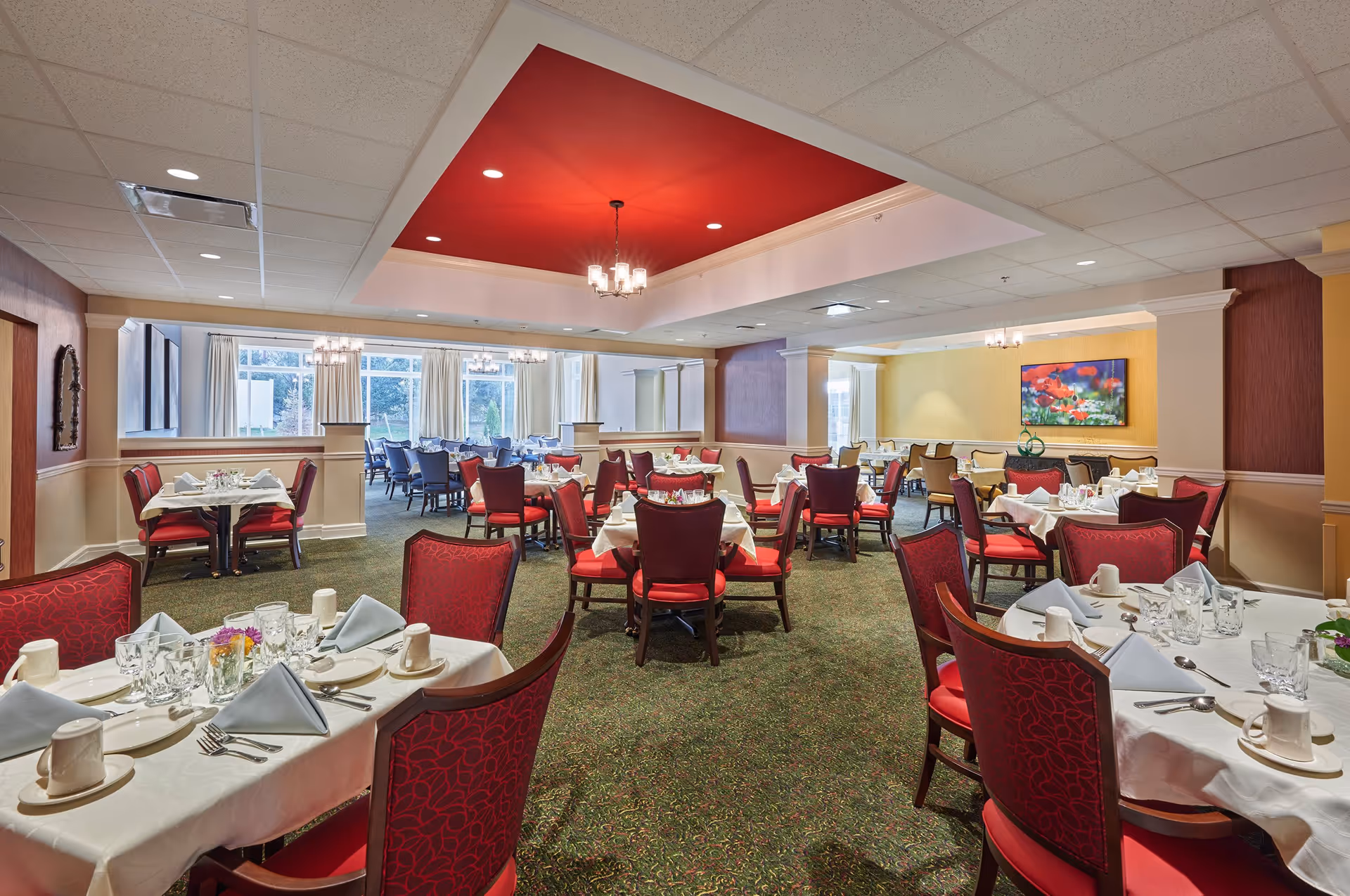 Spacious dining room with neatly set tables, red upholstered chairs, chandeliers, and large windows.