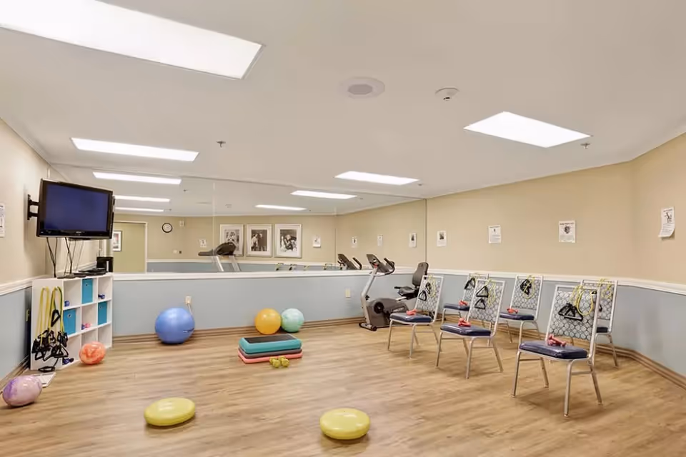 Exercise/activity room with chairs, exercise balls, step platforms, resistance bands, a stationary bike, wall mirror and TV.