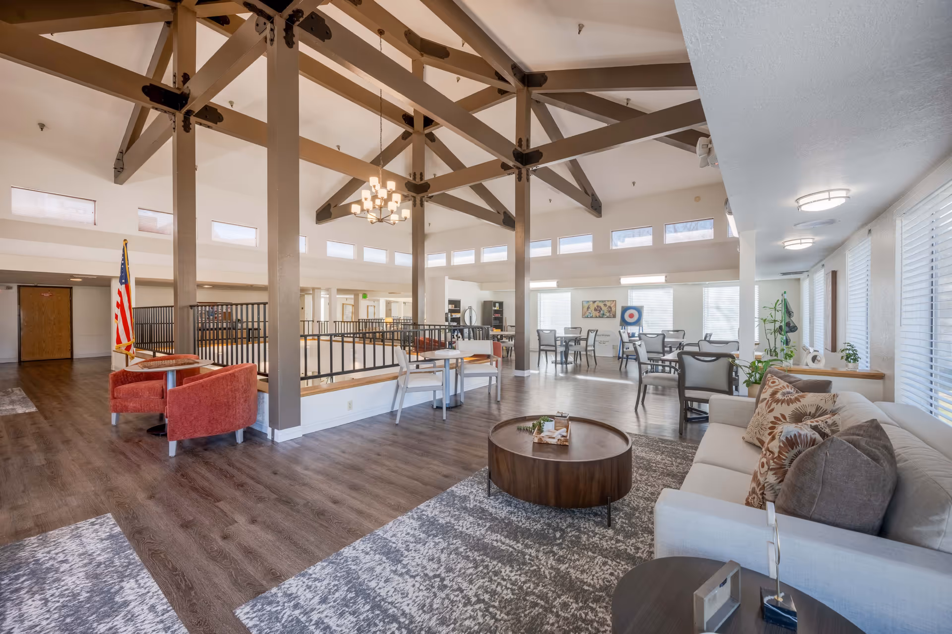 Spacious senior living facility common area with high vaulted ceilings supported by exposed wooden beams. The room features a seating area with a white couch adorned with patterned pillows, a round wooden coffee table on a textured rug, and several tables and chairs arranged for socializing or activities. Large windows with blinds allow natural light to fill the space. An American flag stands near a small round table with two red upholstered chairs. The flooring is wood laminate, and the walls are painted white, creating a bright and welcoming atmosphere.