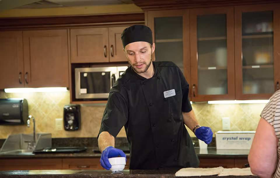 A male chef wearing a black uniform, black hat, and blue gloves is placing a small white bowl on a kitchen counter. The kitchen has wooden cabinets, a microwave, and a sink in the background. A person in a striped shirt is partially visible on the right side of the image.