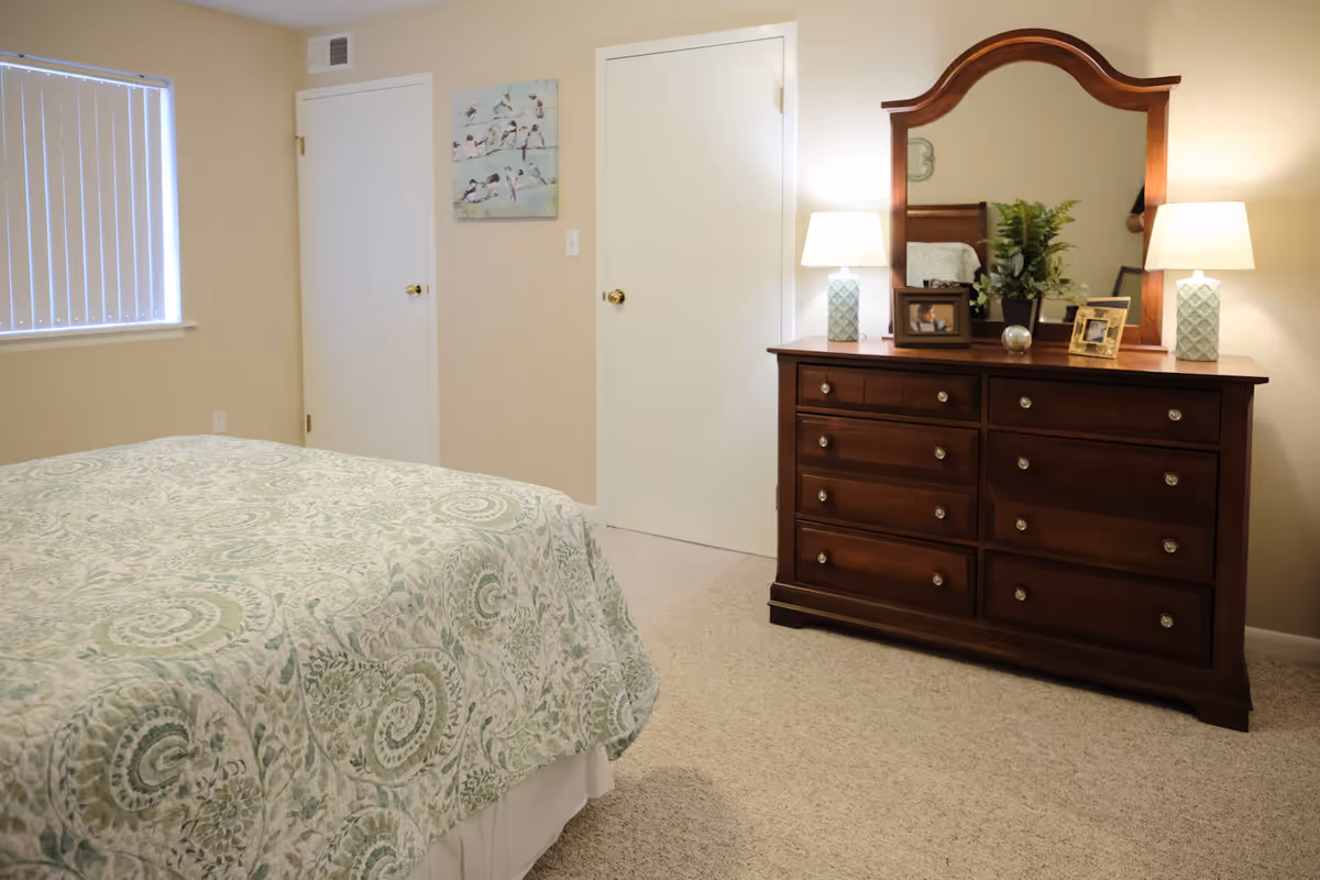 A bedroom with a bed covered in a green and white patterned quilt, a wooden dresser with a large mirror, two table lamps, framed photos, and a plant. There are two closed white doors and a window with vertical blinds.