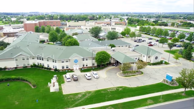 Aerial view of a senior living campus showing connected beige buildings with green roofs, a parking lot, drive-up entrance, and surrounding lawns and trees.