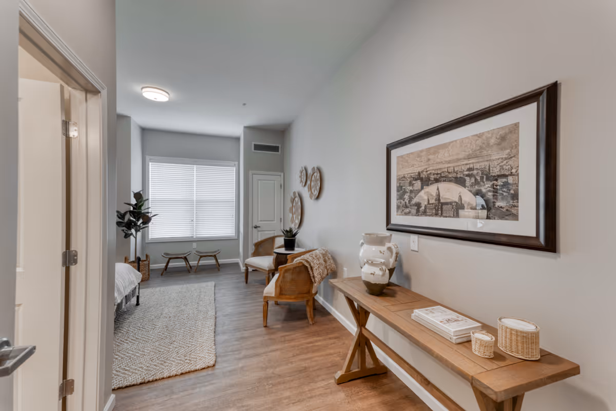 A bright and cozy senior living bedroom with a large window covered by blinds, two wooden chairs with cushions, a wooden bench with decorative items including a vase and books, a framed artwork on the wall, and a soft area rug on the wooden floor.