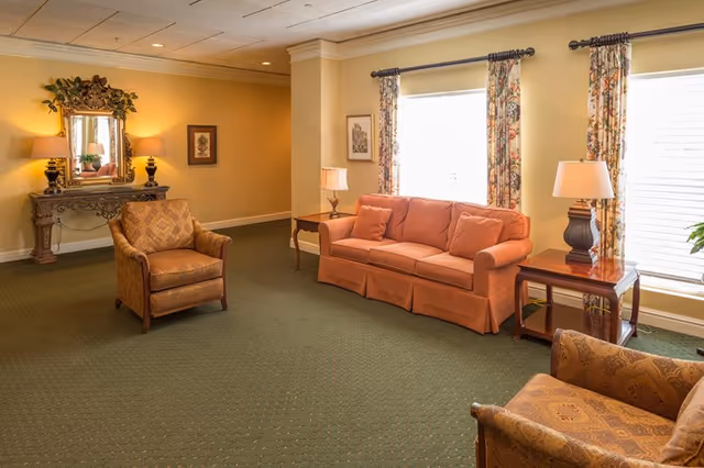 A cozy living room area with a green carpet, an orange sofa with matching pillows, two patterned armchairs, two wooden side tables with lamps, floral curtains on the windows, and a decorative console table with a mirror and lamps against a yellow wall.