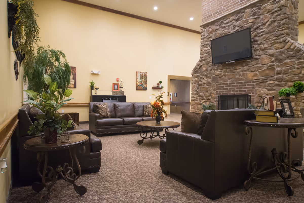 Cozy assisted living lounge with leather sofas, coffee tables, plants, and a stone fireplace with a mounted TV.