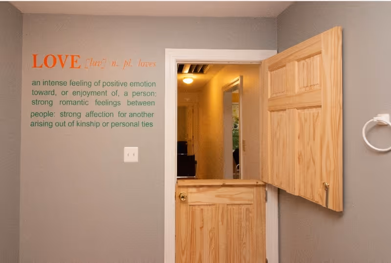 Interior view of a gray-walled room with a wooden Dutch door open to a hallway and a wall decal defining "LOVE".