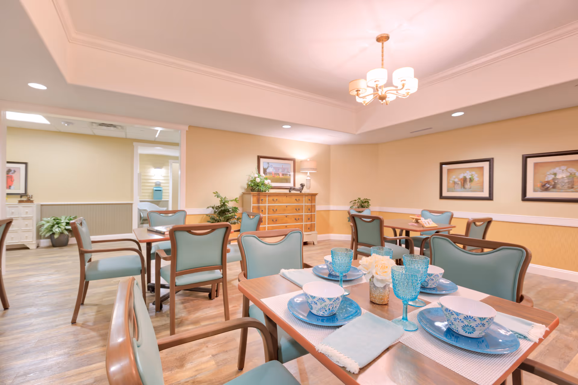 Bright dining room with teal-upholstered chairs, set tables, a chandelier, and framed artwork in a senior living facility.