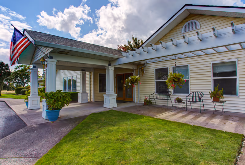 Exterior view of a single-story senior living facility with beige siding, a covered entrance supported by white columns, hanging flower pots, black metal chairs, and an American flag mounted near the entrance. The sky is partly cloudy and there is a well-maintained lawn in front.
