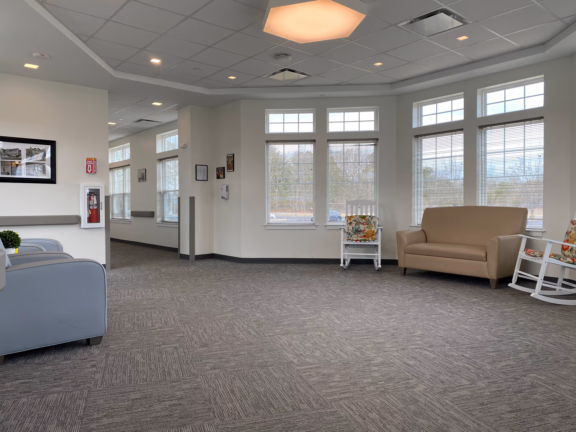A bright and spacious senior living facility common area with large windows letting in natural light. The room features a beige loveseat, two white rocking chairs with floral cushions, and a gray armchair. The floor is carpeted in a gray pattern, and the ceiling has a modern light fixture. There are framed pictures on the walls and a fire extinguisher mounted near the entrance.