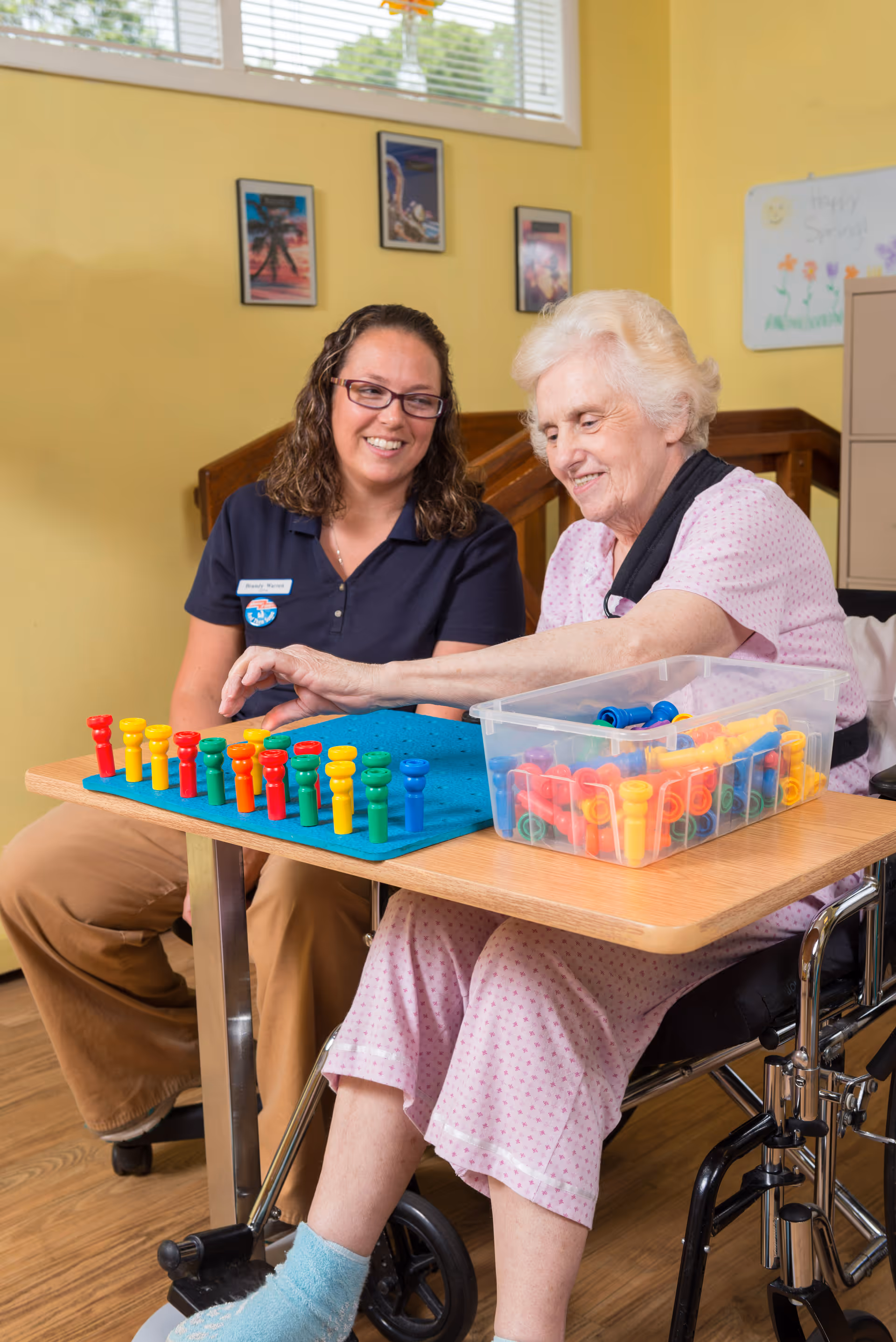 An elderly woman in a wheelchair playing a peg board game with a caregiver in a room with yellow walls and framed pictures. The caregiver is smiling and watching the woman as she places colorful pegs on the board.