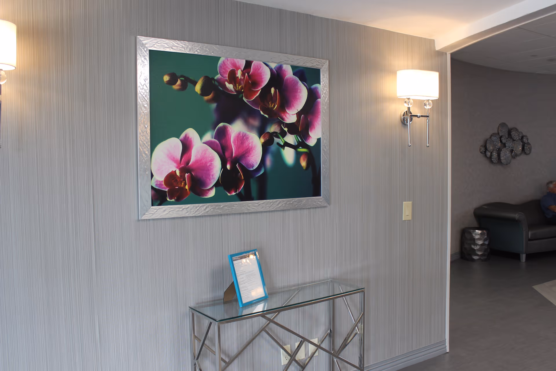 Lobby interior with a large framed orchid print above a glass console table and wall sconces, with seating visible in an adjacent area.