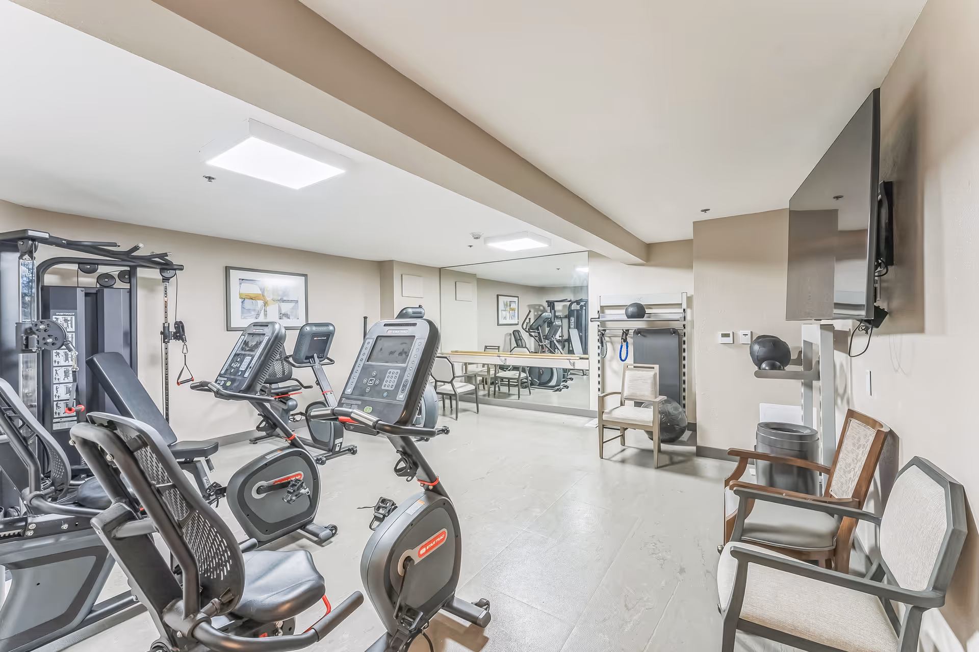 A fitness room with exercise equipment including stationary bikes, a multi-function weight machine, and a rack with exercise balls and other fitness accessories. There are chairs along the wall and a large mirror reflecting the room. A flat-screen TV is mounted on the wall.