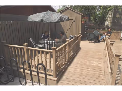 Sunlit wooden outdoor deck with tables, chairs, umbrella, and residents seated outdoors.