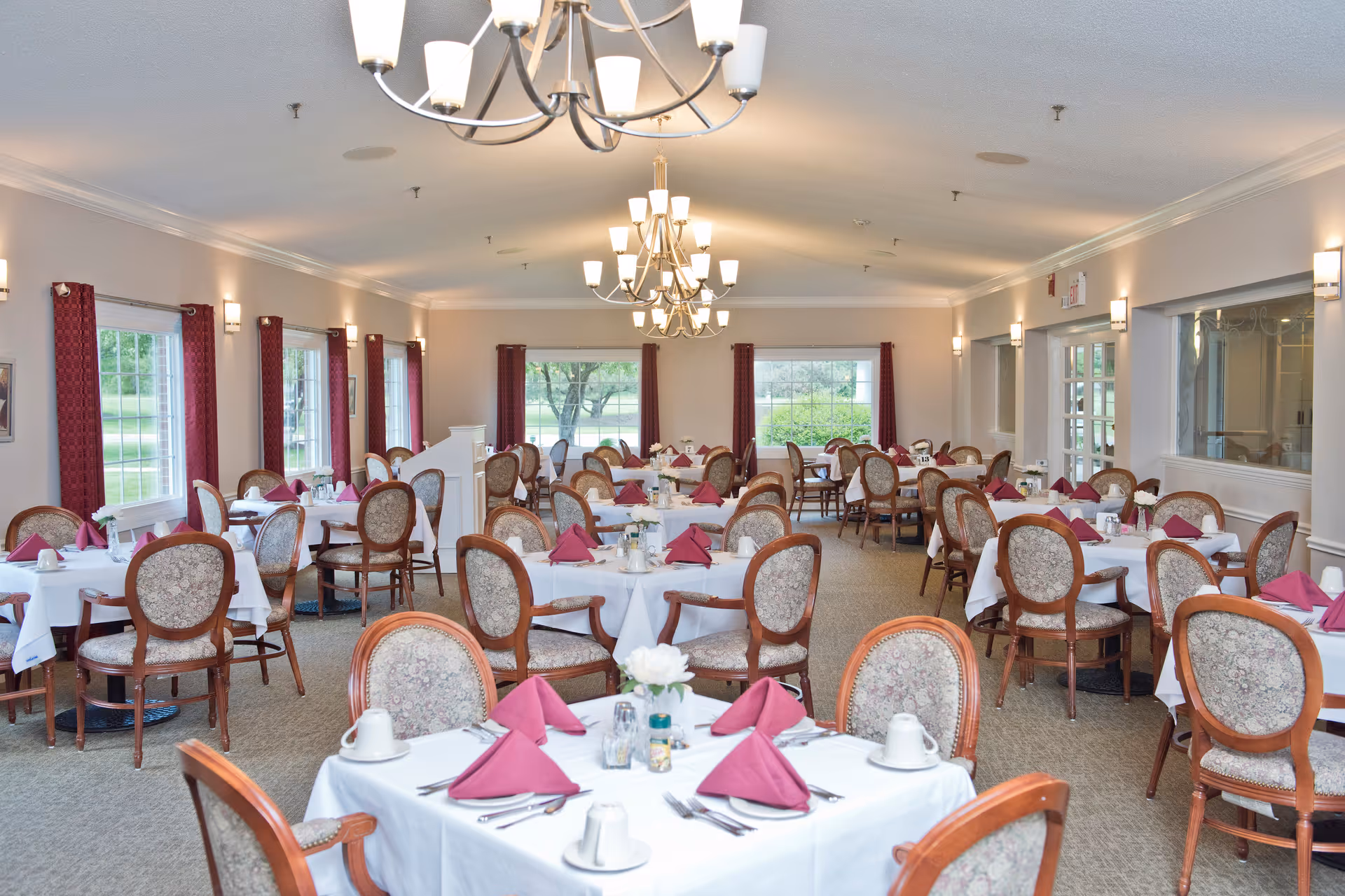 A spacious dining room with multiple round tables covered with white tablecloths, each set with maroon folded napkins, cups, and silverware. The room has large windows with maroon curtains, beige walls, and elegant chandeliers hanging from the ceiling. Upholstered wooden chairs surround the tables.
