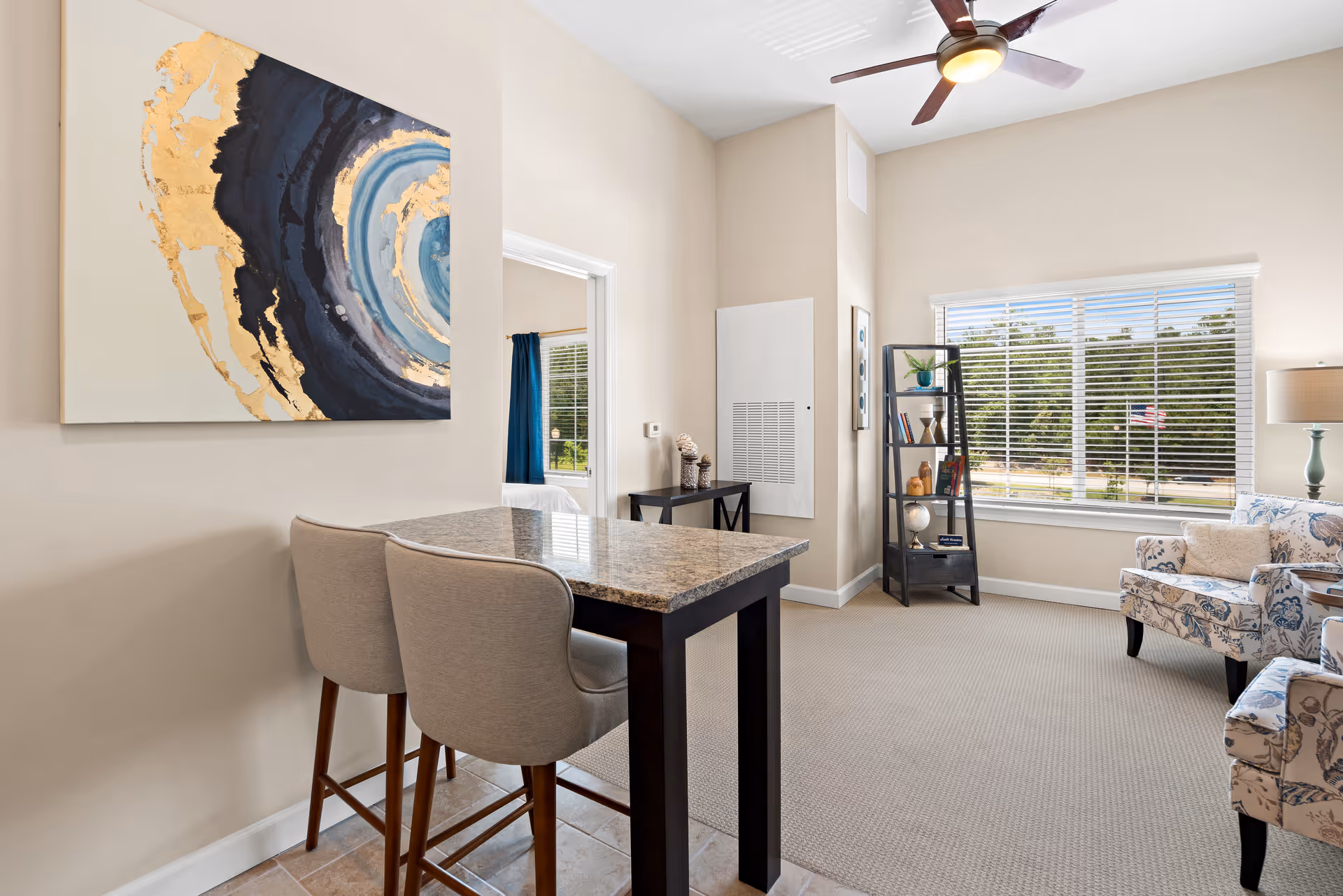 A bright living room area with beige walls and carpet, featuring a granite-topped bar table with two upholstered bar stools in the foreground. A large abstract painting with blue, black, and gold tones hangs on the left wall. In the background, there is a window with white blinds showing an outdoor view with trees and an American flag. The room also includes a ceiling fan with light, a black shelving unit with decorative items, and two floral-patterned armchairs with a side table and lamp.