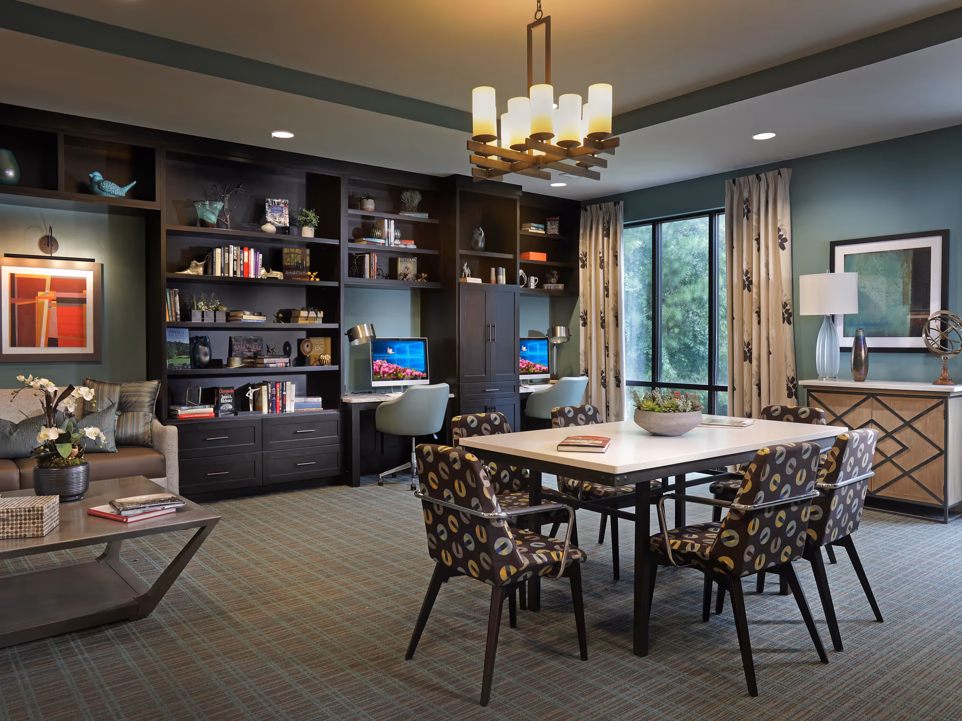 A well-lit common area with a large table surrounded by six patterned chairs in the center. Behind the table, there are two computer desks with chairs and desktop computers, built into a dark wood shelving unit filled with books and decorative items. To the left, there is a cozy seating area with a sofa, a coffee table with books, and a framed artwork on the wall. Large windows with patterned curtains allow natural light to enter the room. A modern chandelier hangs from the ceiling, and a sideboard with a lamp and decorative items is visible on the right.