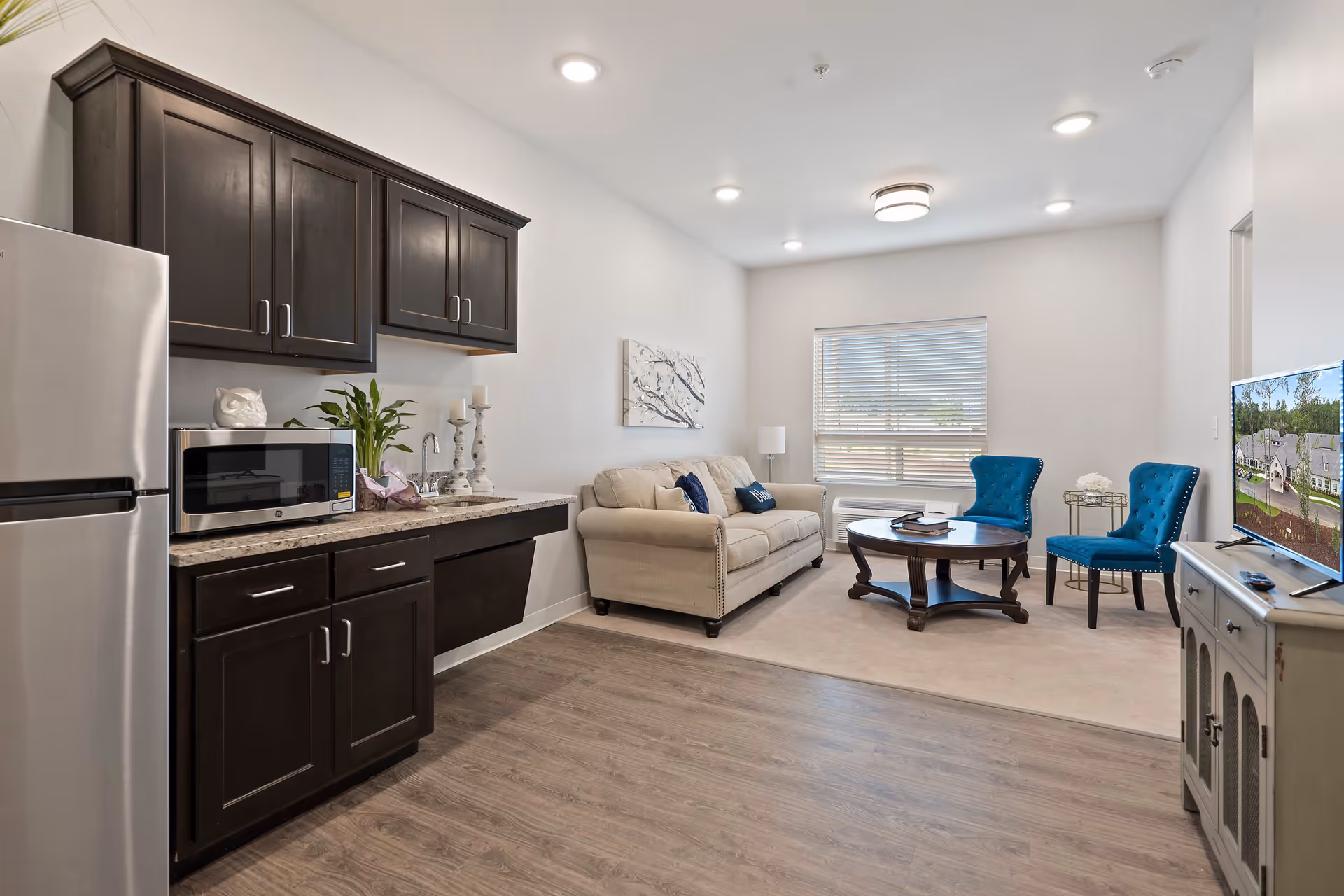 Studio living area with a kitchenette on the left, beige sofa, blue accent chairs, round coffee table and a TV.