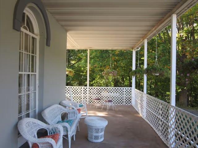 A covered outdoor porch area with white wicker chairs and a matching wicker table. The porch has white lattice railing and hanging flower pots. Trees and greenery are visible beyond the porch.