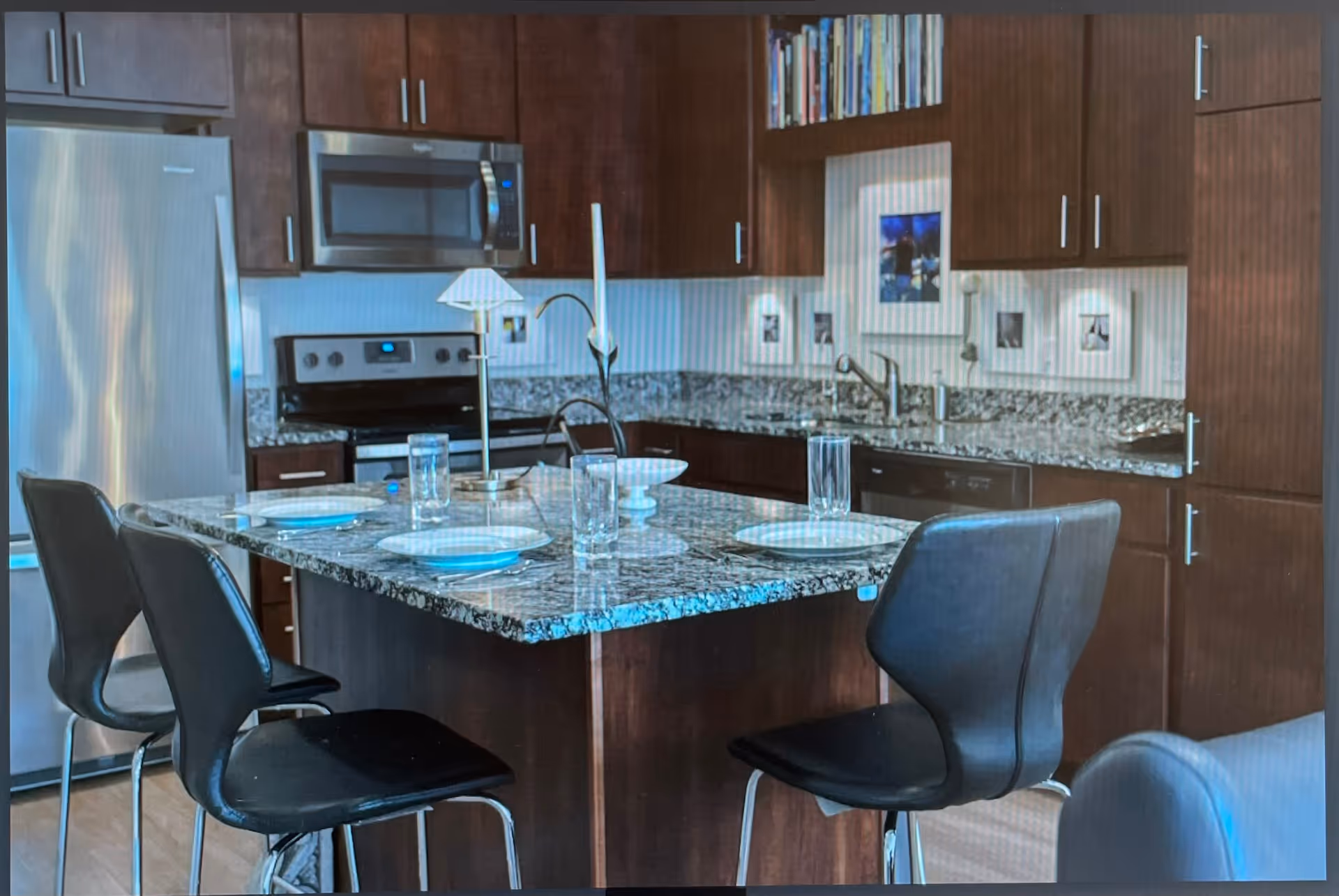 Modern kitchen with dark wood cabinets, stainless steel refrigerator, microwave, stove, and dishwasher. A granite countertop island with four black chairs is set with plates and glasses. Books and framed pictures are displayed on the wall above the sink.