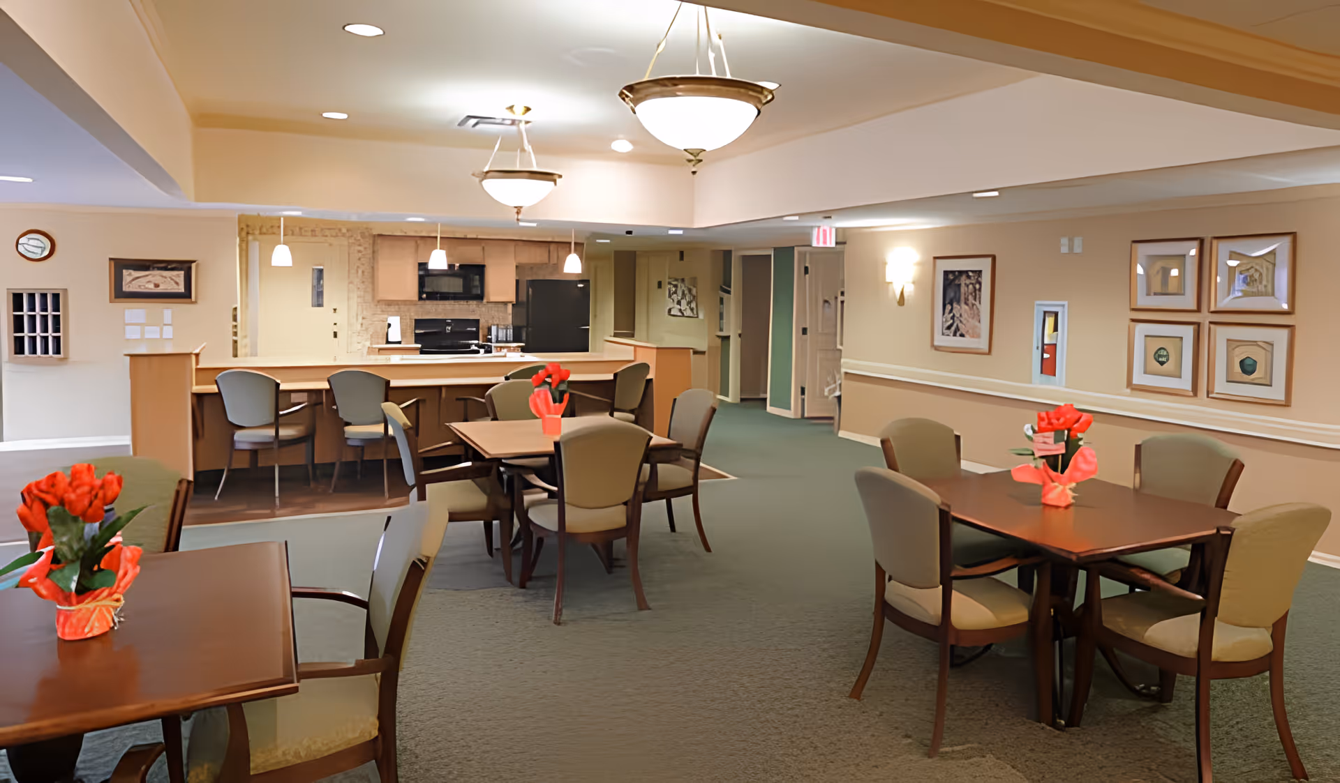 Interior view of a senior living facility dining area with several wooden tables and cushioned chairs arranged around them. Each table has a small red flower centerpiece. In the background, there is a kitchen area with a counter, bar stools, and hanging pendant lights. The walls are decorated with framed artwork and the space is well-lit with ceiling lights.