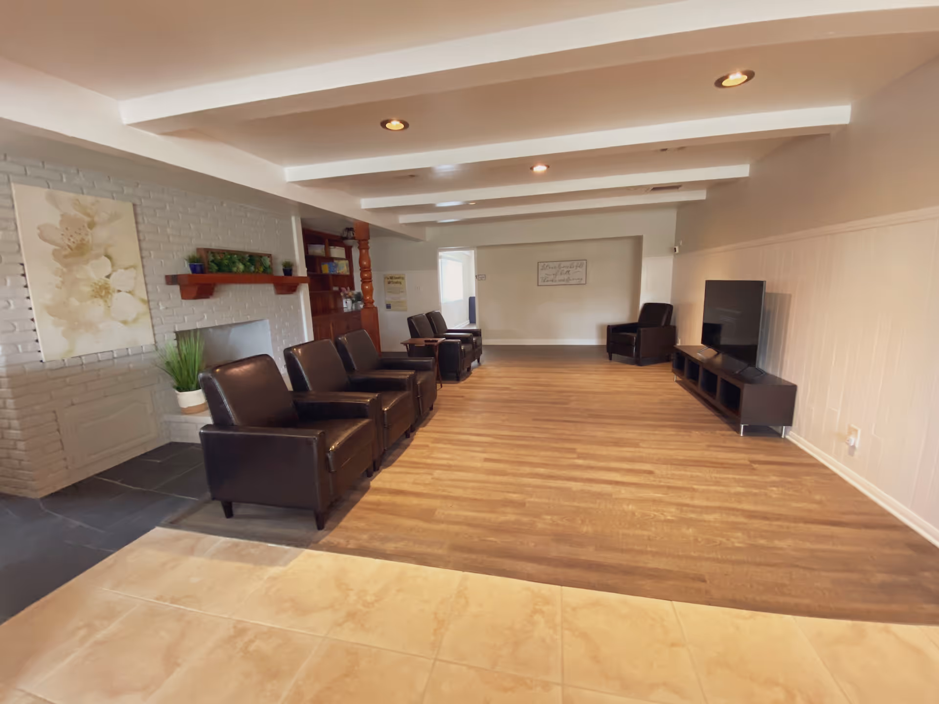 A spacious living room area with wooden flooring and beige tiled entrance. The room features a row of dark brown leather armchairs along the left side near a white brick wall with a floral painting and a wooden shelf with plants. On the right side, there is a flat-screen TV on a low black TV stand. The ceiling has white beams with recessed lighting.