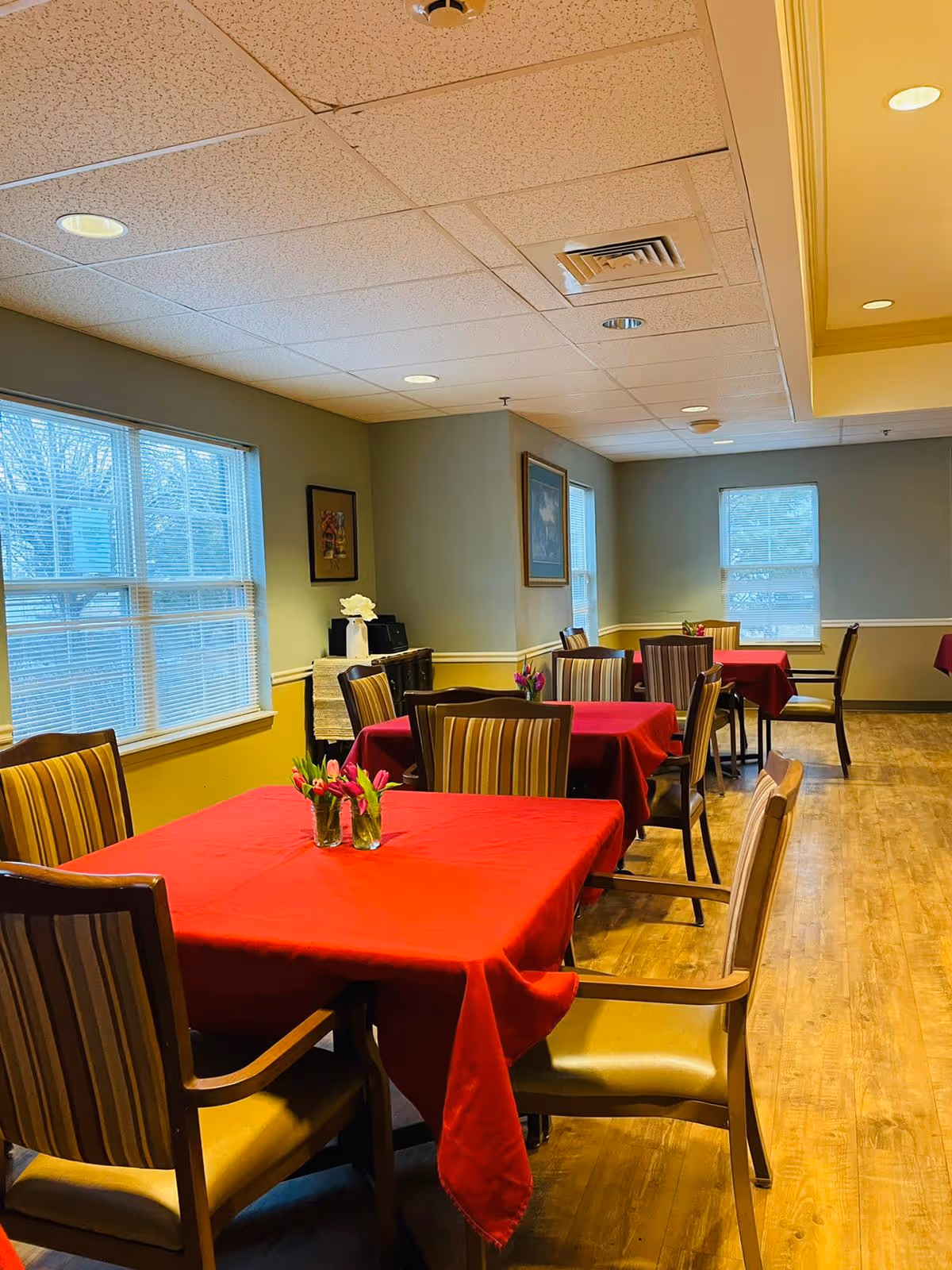 Interior view of a dining room with several tables covered in red tablecloths, each adorned with small vases of flowers. The room has wooden floors, striped cushioned chairs, large windows with blinds, and framed artwork on the walls.