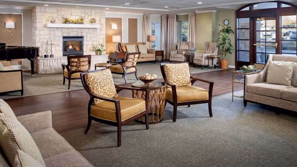 A spacious senior living facility common area with comfortable seating including armchairs and sofas arranged around small tables. There is a stone fireplace with a fire burning, a grand piano to the left, and large windows letting in natural light. The room has a mix of carpet and wood flooring, with lamps and plants adding to the cozy atmosphere.