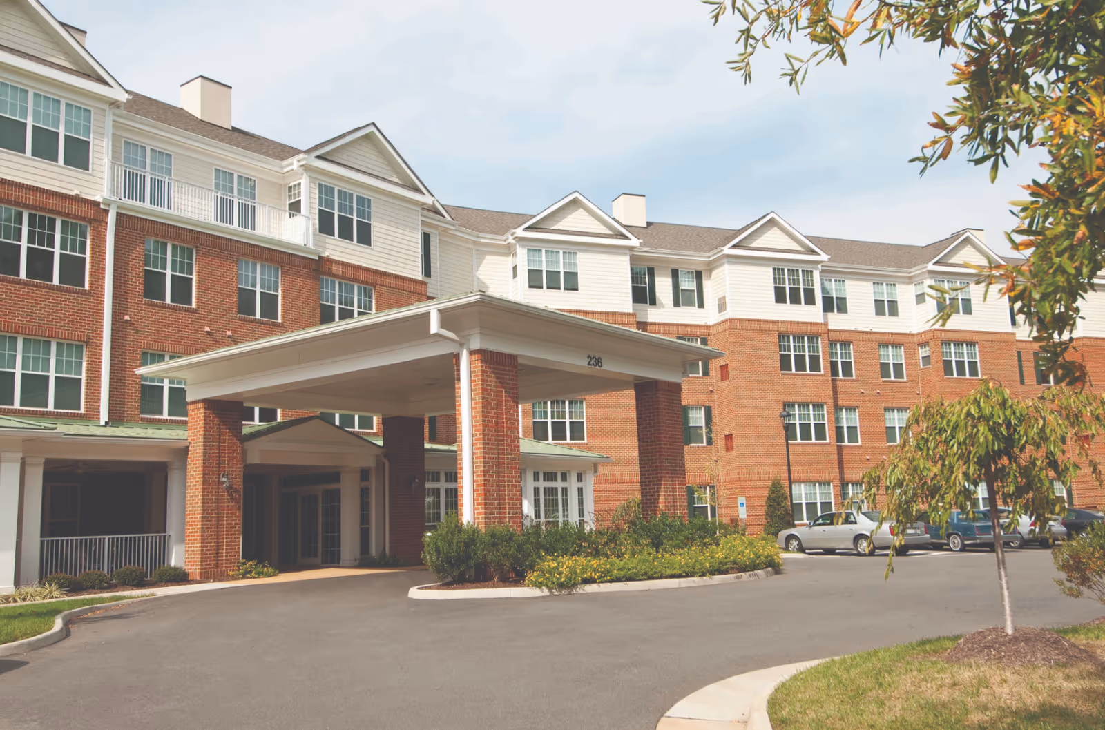 Front entrance of a multi-story brick-and-siding senior living building with a covered porte-cochere and parked cars.